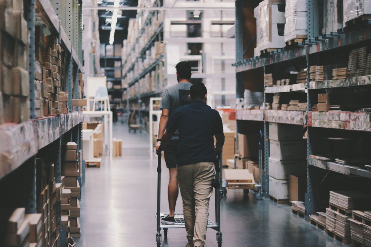 Home Two men maneuver a trolley in a large warehouse filled with boxes and shelves.